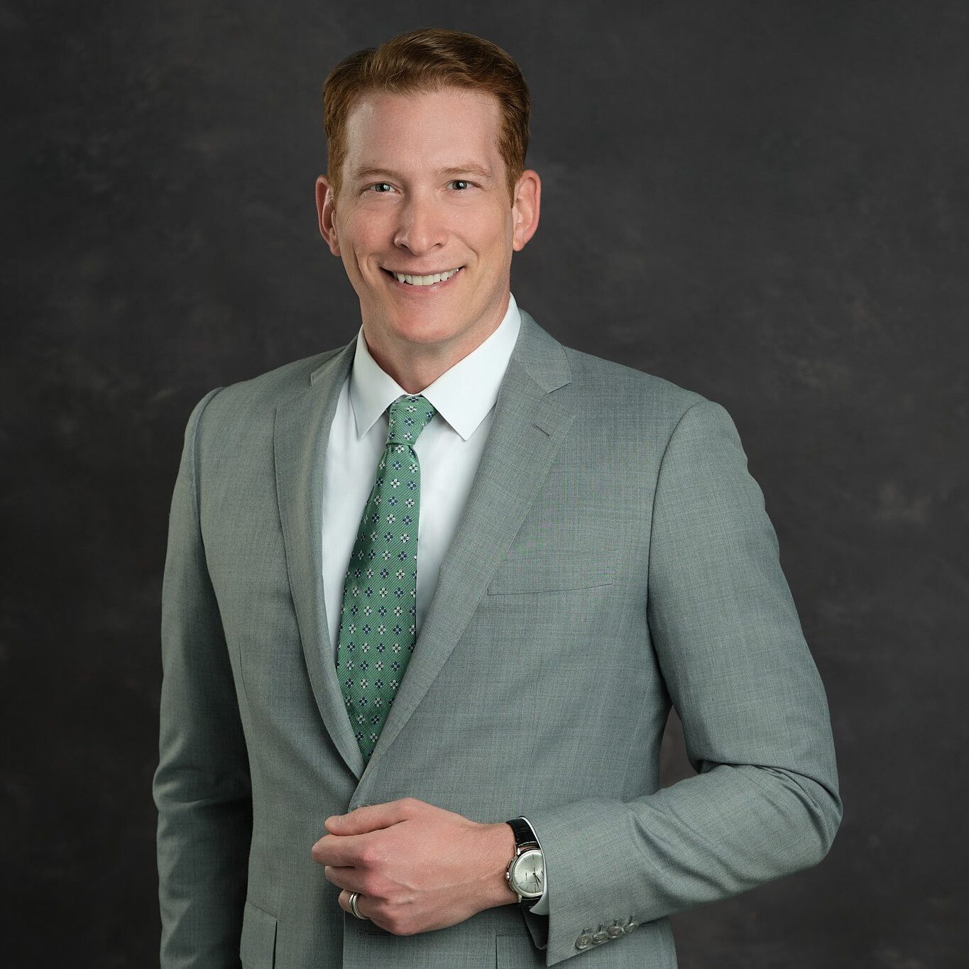 Portrait of Dr. Daniel Parker, smiling and dressed in a light gray suit with a white shirt and green tie, standing against a dark background.