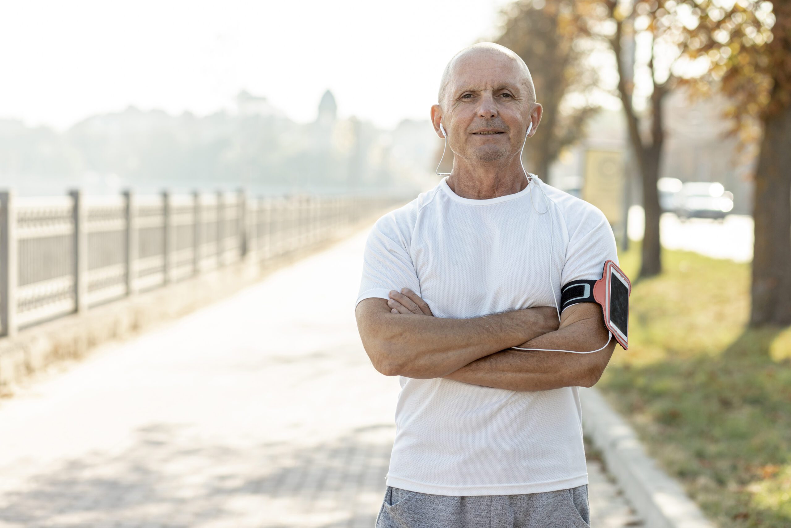 An older man in athletic wear and earphones stands confidently with arms crossed on a sunny path, ready for a run.